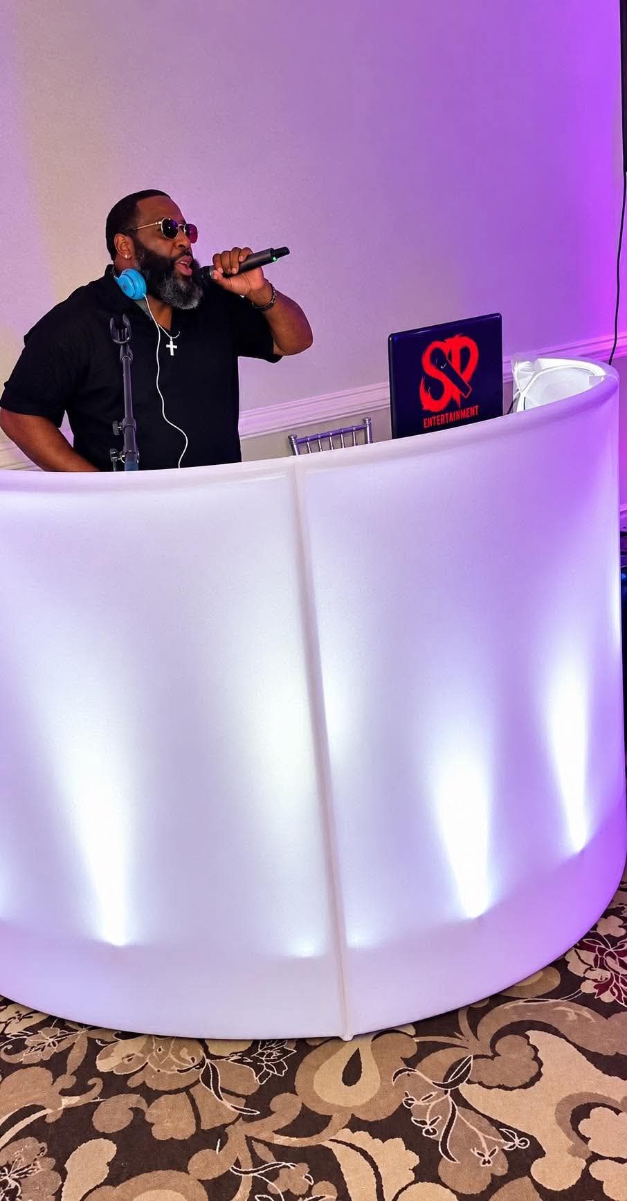 DJ Stevie Steve speaking on a wireless microphone beside an illuminated white DJ booth with an SP Entertainment laptop