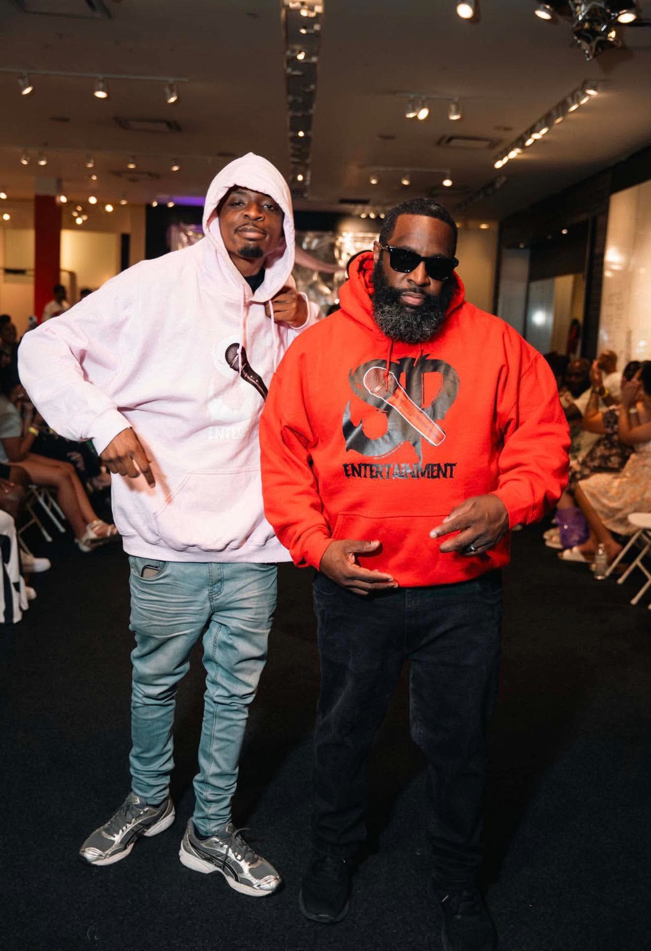 Two men posing at an event, one in a pink hoodie and one in a red SP Entertainment hoodie, guests seated along a runway behind them